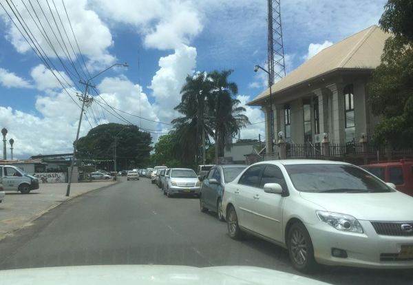 Lange rij auto's in Surinaams straatbeeld met palmen, electriciteitsleidingen en erboven een wolkenlucht